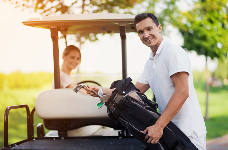 Smiling man pulling golf club from bag beside golf cart with woman seated