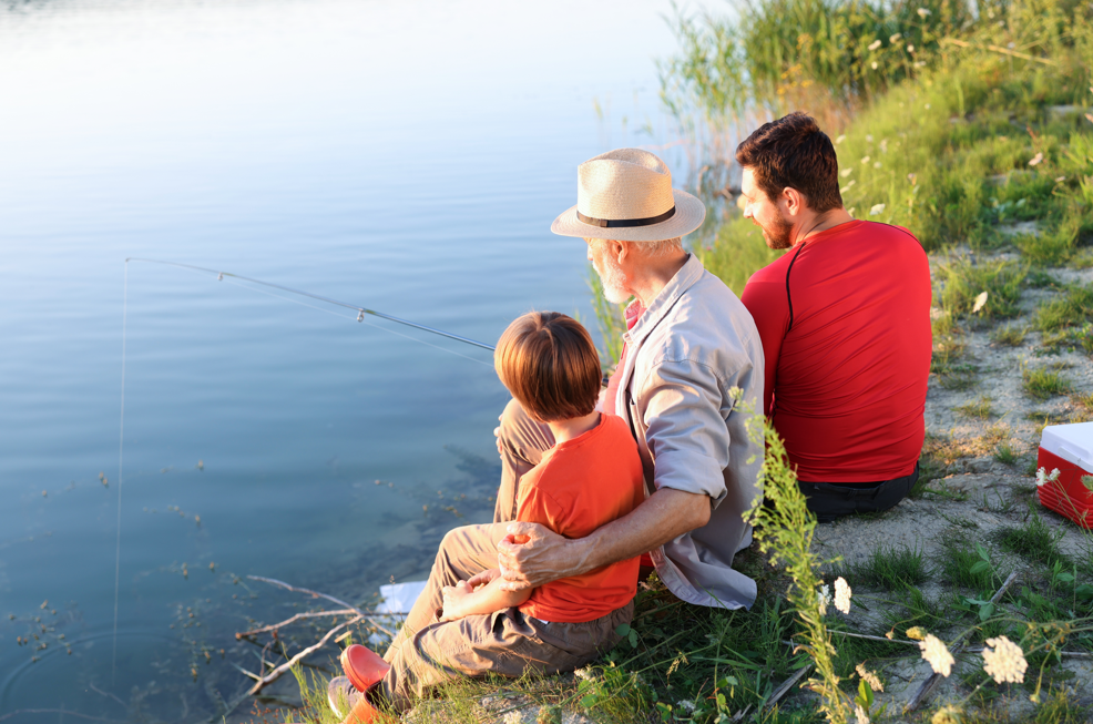 Grandfather, father, and son fishing together from grassy lakeshore at sunset